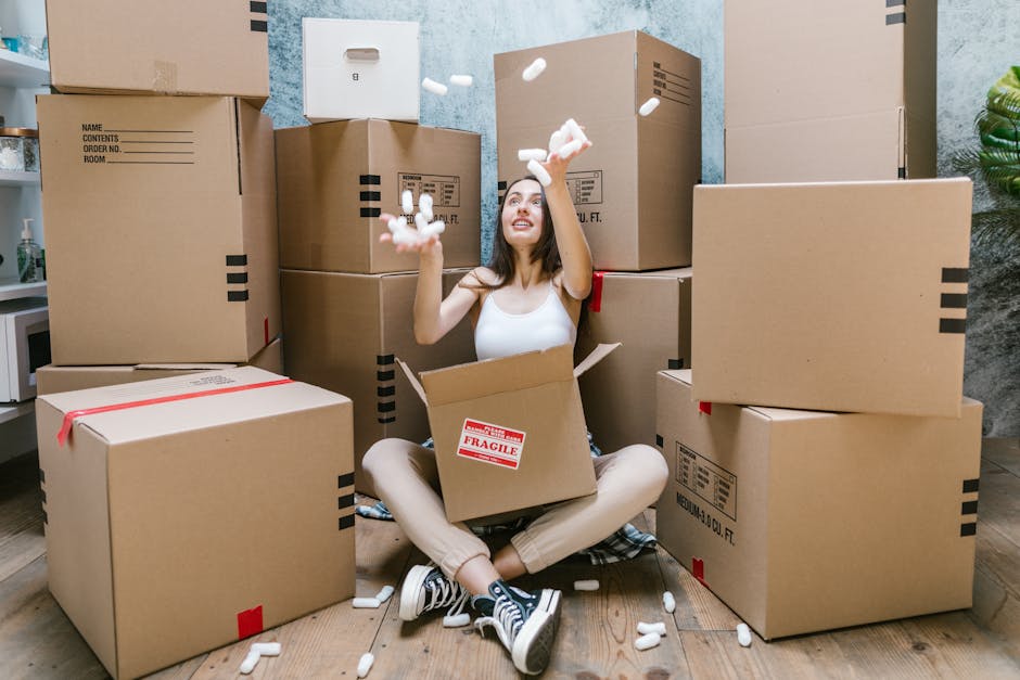 A young woman sitting on a wooden floor surrounded by stacked cardboard packing boxes during a home relocation, with some boxes sealed with red tape and labelled, while she throws packing foam peanuts into the air. She is inside a room with a textured blue wall and a small portion of a kitchen visible on the left side, including a microwave and potted plant. The boxes vary in size and are arranged both on the floor and stacked against the wall, indicating an active packing or unpacking process. The woman is dressed casually in a white tank top, beige pants, and sneakers, suggesting a move or packing day. This scene illustrates furniture transport and packing activities typical of house removals, aligning with services provided by Kennington Removals for efficient relocation within London.