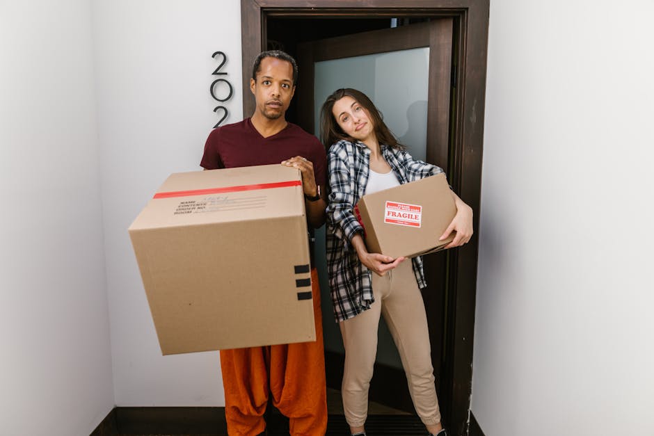 A man and a woman standing in a doorway inside a residence, each holding cardboard boxes used for packing and moving. The man, on the left, is wearing an orange-brown pair of trousers and a maroon t-shirt, with a serious expression, while holding a large box with black tape securing it. The woman, on the right, dressed in beige trousers and a black-and-white checkered shirt, has a slight smile and is holding a smaller box marked with a red 'fragile' label. They are positioned close together in front of a dark wooden door frame, with a white wall and a numbered sign beside them indicating the property number. Inside the doorway, there are packing materials such as brown cardboard boxes, and the ambient lighting suggests an indoor environment prepared for a home relocation. This scene illustrates the process of furniture transport, packing, and loading during a house removal with the assistance of Kennington Removals, specializing in same-day relocation services in the Kennington SE11 area.