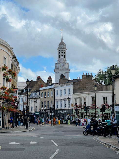 A busy street scene in Kennington showing a mix of pedestrians walking along the pavement and crossing the road, with some individuals carrying parcels or pushing strollers. Classic terraced buildings with shopfronts and hanging flower baskets line the street, and a vintage-style lamppost is visible on the corner. In the background, a tall historic clock tower with a spire topped by a weather vane rises above the surrounding rooftops. A white van with the branding of Kennington Removals is parked nearby, and a few people are loading or unloading furniture and boxes into the vehicle. The sky is partly cloudy, providing natural light that illuminates the urban environment. This scene illustrates a typical setting for home relocation and furniture transport, with ongoing activities related to packing, moving, and street-based logistics in the context of house removals in Kennington, as described on the company's website.
