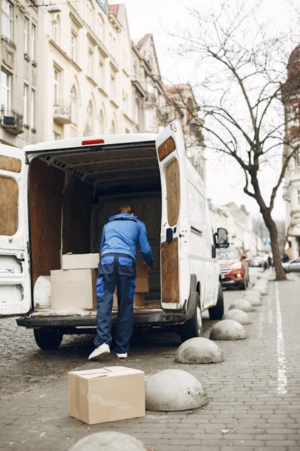 A man wearing a blue jacket and dark trousers is seen loading or unloading cardboard boxes into the back of a white moving van parked on a city street with a cobblestone sidewalk. The van's rear doors are wide open, revealing an empty interior with wooden paneling, suitable for furniture transport and packing during a home relocation. Several cardboard boxes, some on the ground and one inside the van, are visible, indicating packing activity. The urban setting features multi-story period-style buildings with ornate facades, leafless trees lining the street, and large rounded concrete bollards along the pavement. The scene suggests a professional removals process by Kennington Removals, supporting efficient furniture transport and moving logistics in a residential area during daylight.