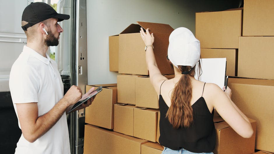 A man and a woman are inside a property during a house removal process, with the man holding a clipboard and a pen, and the woman wearing a white cap and black top, reaching up to check or move stacked cardboard boxes on a wall-mounted shelving unit. The boxes are arranged in several stacks and appear to contain household items prepared for transport. The environment is well-lit, with natural light coming from an open door or window nearby, and the scene depicts the packing and loading stage of a home relocation. Kennington Removals staff perform tasks such as organizing and preparing belongings for safe furniture transport, involving the use of packing materials, including cardboard boxes, to facilitate efficient packing and moving services.