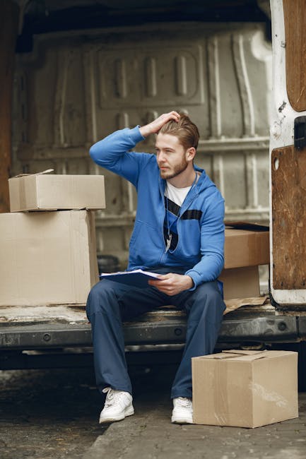 A young man with light brown hair and a beard, dressed in a blue zip-up hoodie, dark trousers, and white sneakers, is sitting on the edge of a moving van's open cargo area. He is holding a clipboard in his left hand and appears to be contemplating or taking a break during a home relocation process. Several cardboard boxes of varying sizes, some sealed with packing tape and others open, are placed around him inside and outside the van. The interior of the van shows visible wooden panels and a metal floor, with a partially visible door on the right side. The moving vehicle is parked outdoors, on a paved surface near a building, and the scene is lit with natural daylight. This image relates to furniture transport and packing as part of a professional removals service, illustrating the loading process managed by Kennington Removals during house moves.