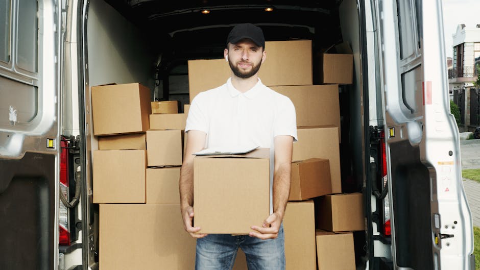 A white moving van parked on the street in front of a brick building with large windows, with the driver visible in the cab. The van is labeled 'MOVING COMPANY' with smaller text 'LOCAL & LONG DISTANC