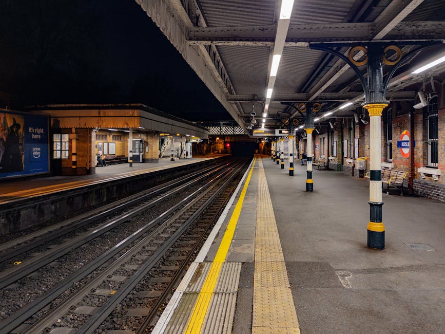 A nighttime view of a London Underground train station platform with several yellow and black safety poles lined along the edge, a tactile paving strip, and a row of empty benches against the brick walls. The platform is illuminated by overhead lighting, with tracks running parallel to the platform and extending into the distance. The station's canopy, supported by decorative metal columns with yellow accents, covers the platform area, while platforms on the opposite side are visible with a person sitting on a bench near a small ticket booth. This setting depicts a typical urban rail station environment suitable for house removals and furniture transport logistics, with clear pathways for loading and unloading goods. Visual cues such as the trolleys and packaging materials are not present, but the scene captures the essential features of a transport hub relevant to professional removals services.