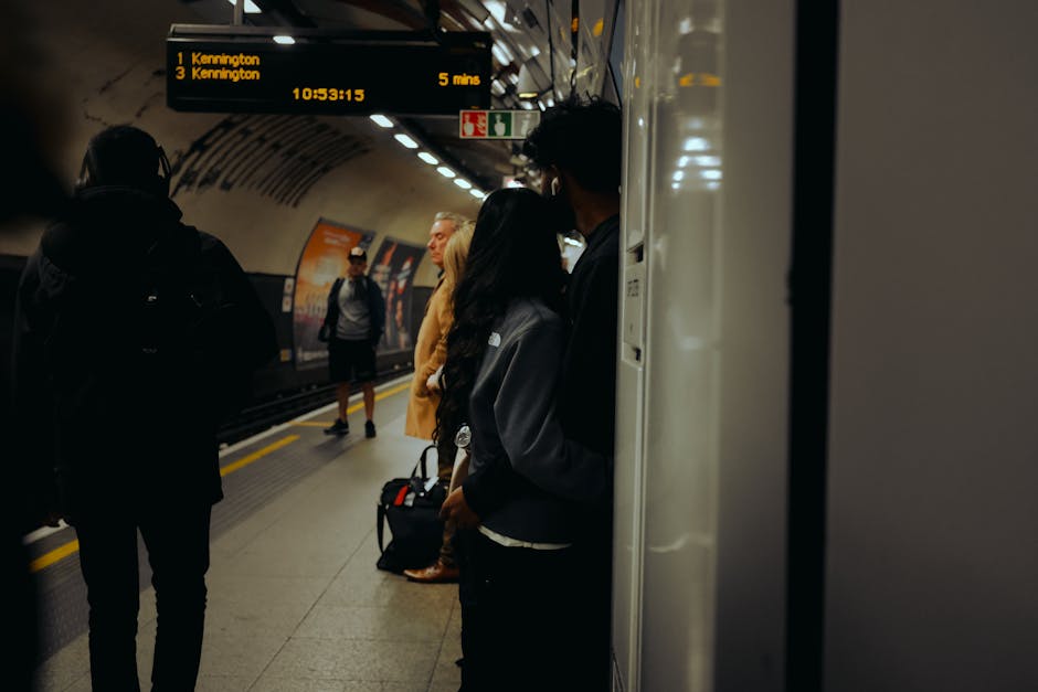 Inside an underground train station, several passengers are waiting on the platform near the edge, prepared for their journey. The platform surface is made of concrete with yellow safety lines, and the station walls are lined with advertising posters. Overhead, an electronic departure board displays train information, including the next train to Kennington departing from platform 1 in five minutes, with the current time shown as 10:53:15. Some passengers, wearing jackets and carrying backpacks or rolling suitcases, are standing close to the platform edge, while others are further back. One individual is partially visible, leaning against a column or wall, with their face turned away, and holding a bag. The lighting in the station is bright but ambient, creating a neutral and functional atmosphere typical of city transit systems. This setting is relevant to house removals or relocation services, as it illustrates the travel and transport environment associated with home or office moves, similar to those managed by Kennington Removals, especially when involving moving logistics and transportation planning.