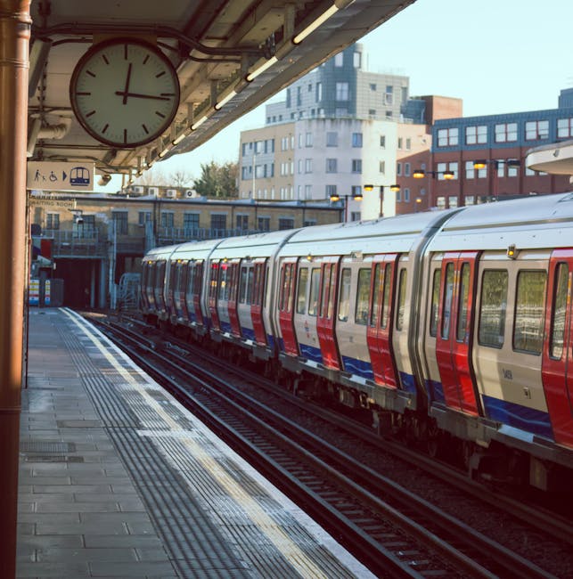 A nighttime view of a London Underground train station platform with several yellow and black safety poles lined along the edge, a tactile paving strip, and a row of empty benches against the brick walls. The platform is illuminated by overhead lighting, with tracks running parallel to the platform and extending into the distance. The station's canopy, supported by decorative metal columns with yellow accents, covers the platform area, while platforms on the opposite side are visible with a person sitting on a bench near a small ticket booth. This setting depicts a typical urban rail station environment suitable for house removals and furniture transport logistics, with clear pathways for loading and unloading goods. Visual cues such as the trolleys and packaging materials are not present, but the scene captures the essential features of a transport hub relevant to professional removals services.