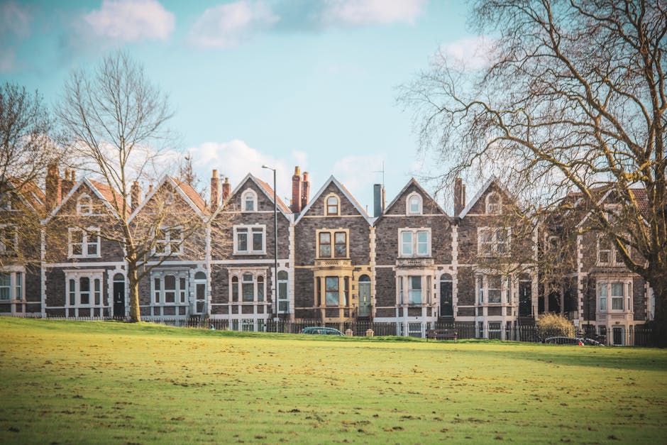 A row of historic Victorian-style terraced houses built from grey stone with white window frames and pitched roofs, situated behind a grassy park area with several leafless trees. The houses are lined up with consistent architectural features, including chimneys and decorative window hoods. In the foreground, the park has a wide, well-maintained lawn, and a paved pathway runs along the front of the houses. The clear sky with a few clouds and the shadows cast by the trees indicate daytime. This image reflects a typical residential setting where a home relocation or furniture transport involving house removals could take place, supported by the professional services of Kennington Removals in their house and apartment moving operations.