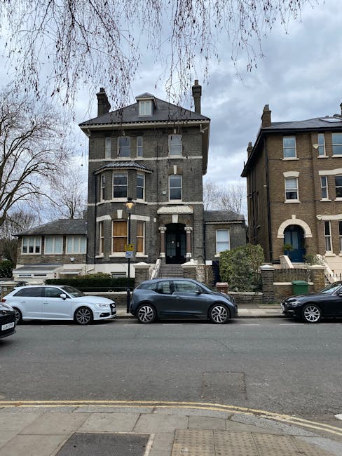 View of a multi-storey Victorian-style house with a bay window and a central entrance, situated on a residential street. Several small trees with bare branches are visible in front of the building. Parked along the street are a white sports car, a dark grey hatchback, and other vehicles. The scene is overcast with grey clouds. Inside the property, a moving company, Kennington Removals, has arranged cardboard boxes, protective wrapping materials, and moving blankets near the entrance, indicating active packing or furniture transport as part of a home relocation or house removal process. The boxes and packaging materials are placed on the pavement and inside the doorway, suggesting preparation for loading into a removal van parked out of view. The environment reflects an organized moving operation, with the focus on furniture transport and relocation logistics, captured during a daylight period.