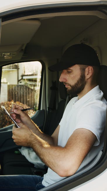 A man wearing a black cap and white T-shirt is seated inside a vehicle, likely a removal van or transporter, during a home relocation process. He is holding a smartphone in his left hand and using a s