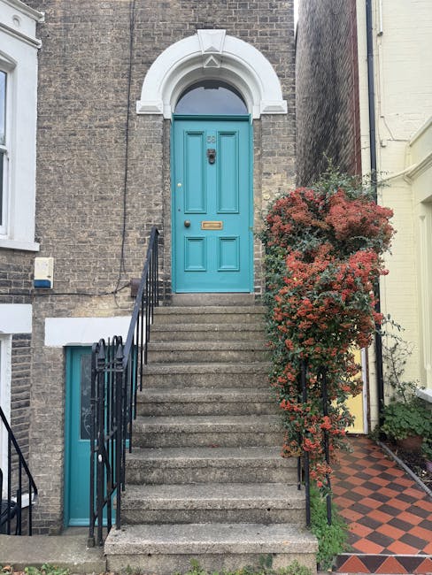 A set of concrete stairs leading up to a bright blue front door in a residential property with a white decorative arch above and a brass mailbox slot. Black metal railings run along both sides of the stairs, with one side featuring a small low-level window with a white frame beneath. To the right of the stairs, there is a tall, bushy plant with reddish-green leaves growing against the brick wall. The property’s facade is made of brown brick, and the adjacent ground is paved with red and black checkered tiles. The image appears to be taken during daylight hours, providing natural lighting that highlights the texture of the brickwork, the details of the orange and red foliage, and the painted door. This scene illustrates a typical home entrance, relevant to house removals and relocation services offered by Kennington Removals, especially when planning for tight access or furniture transport through narrow staircases.