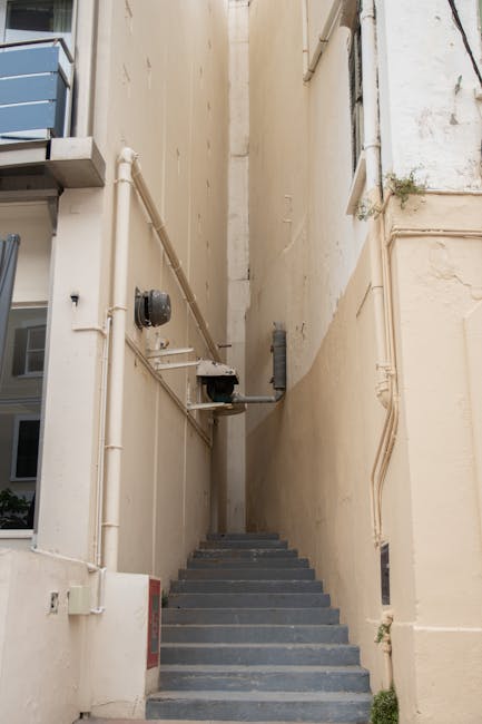 A narrow outdoor staircase with dark grey steps ascending between two beige building walls. The wall on the left has visible piping and a small external electrical box, while the right wall shows some minor weathering and a small plant growing near the bottom. The staircase is used for home relocation access, possibly during a property move or furniture transport, with no vehicles or moving equipment visible in the image. The space is lit by natural daylight, emphasizing the confined nature of the stairway, which presents a challenge for moving large or bulky furniture typical of house removals. Kennington Removals, specialists in removals, may advise on navigating such tight access points for successful relocation logistics.