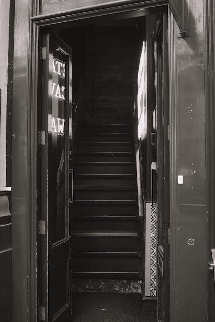 A set of concrete stairs leading up to a bright blue front door in a residential property with a white decorative arch above and a brass mailbox slot. Black metal railings run along both sides of the stairs, with one side featuring a small low-level window with a white frame beneath. To the right of the stairs, there is a tall, bushy plant with reddish-green leaves growing against the brick wall. The property’s facade is made of brown brick, and the adjacent ground is paved with red and black checkered tiles. The image appears to be taken during daylight hours, providing natural lighting that highlights the texture of the brickwork, the details of the orange and red foliage, and the painted door. This scene illustrates a typical home entrance, relevant to house removals and relocation services offered by Kennington Removals, especially when planning for tight access or furniture transport through narrow staircases.
