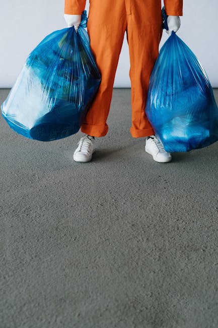 A large pile of mixed household waste and discarded items, including broken appliances, plastic and metal parts, cardboard boxes, and damaged furniture, is situated outdoors on a gravel surface next t