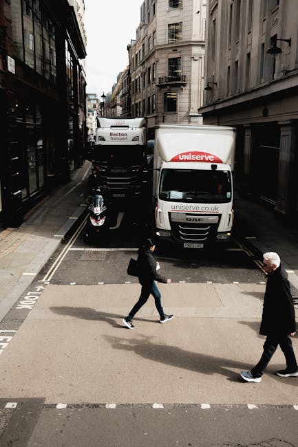 A narrow urban street scene showing a commercial vehicle from Uniserve positioned at a loading area with its rear doors facing a building entrance, indicating a home removal or furniture transport process. The vehicle is parked parallel to the sidewalk, with a motorcycle next to it and a white van in the background. Two pedestrians, a woman carrying a bag and a man, are crossing the street at a pedestrian crossing in front of the vehicle. The surrounding environment includes multi-storey buildings with traditional architecture, black metal fire escapes, and narrow pavements. Overcast lighting provides even illumination across the scene. This setting illustrates a typical moving logistics environment, involving loading or unloading activities that Kennington Removals might coordinate during a house relocation within the area.