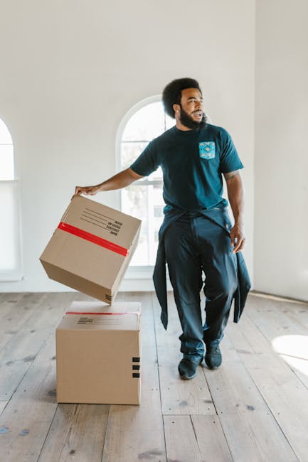 A man with a dark beard and curly hair, dressed in a navy blue t-shirt and trousers, is inside a well-lit room with white walls and wooden flooring, holding a cardboard box with red and black tape. Behind him, there is a large arched window letting in natural light, indicating daytime. The man appears to be in the process of home relocation, possibly preparing for furniture transport or packing and moving tasks. On the floor in front of him are two more cardboard boxes, one stacked on top of the other, both sealed with packing tape. The room is empty apart from the boxes, suggesting the initial stages of a house move or a clearance. This visual aligns with professional removal services offered by companies like Kennington Removals, focusing on house removals, packing, and transport logistics for clients in the SE11 area or during student move-out processes, as highlighted in the page title.